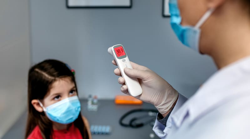 A child getting her temperature checked by a doctor with an infrared themometer, they are both wearing masks