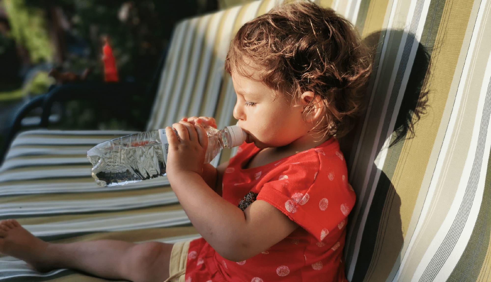 Little toddler girl drinking water from bottle at couch in summer garden