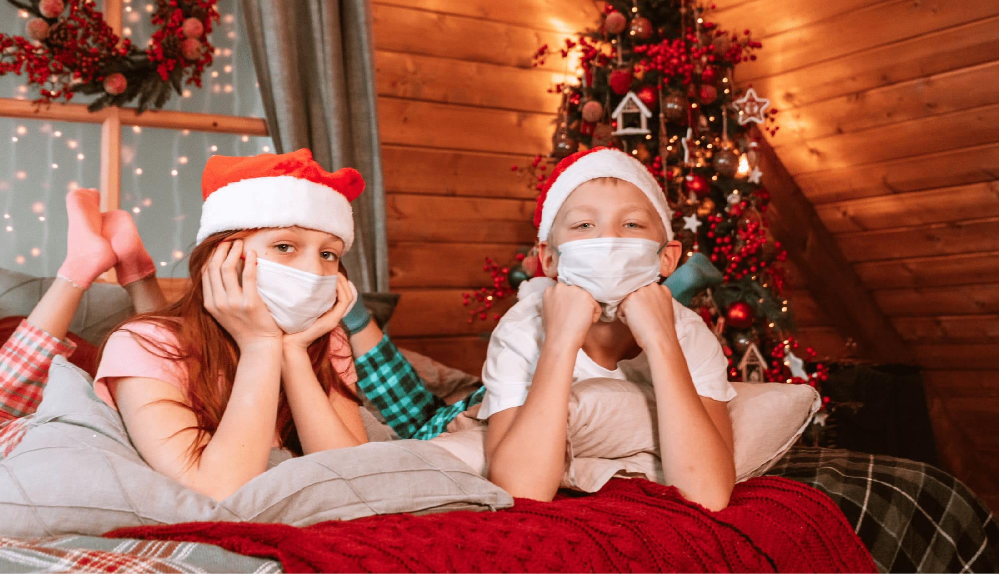 two children boy and girl in pajamas, Santa hats and protective face masks on the bed having fun during the festive season
