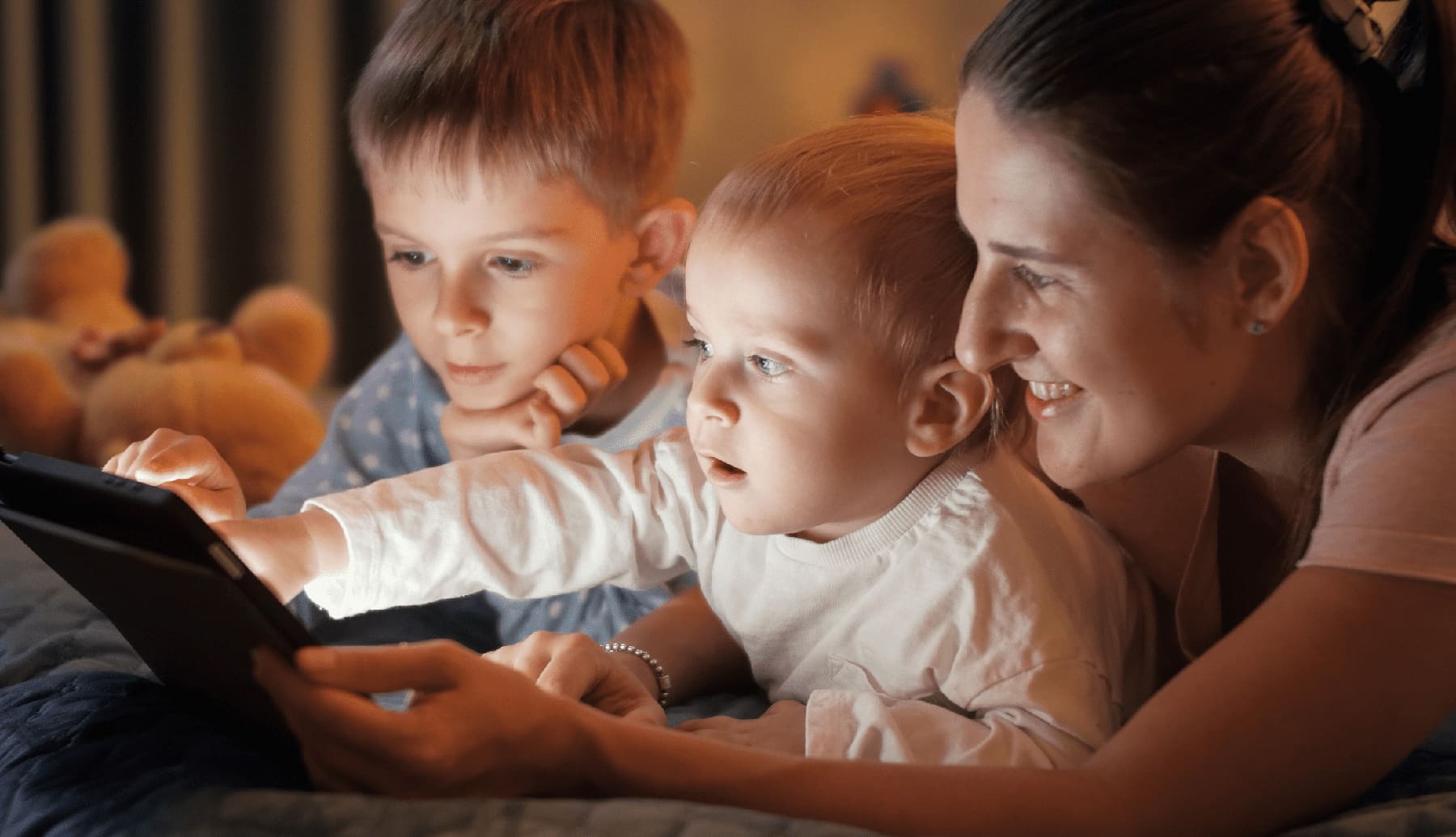 Young mother lying with two boys in bed playing on tablet computer at night