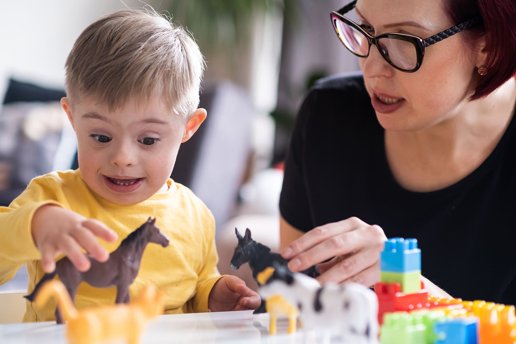 Young child with Down syndrome excitedly playing with animal toys while bonding with a caregiver, highlighting joyful interaction and developmental support.