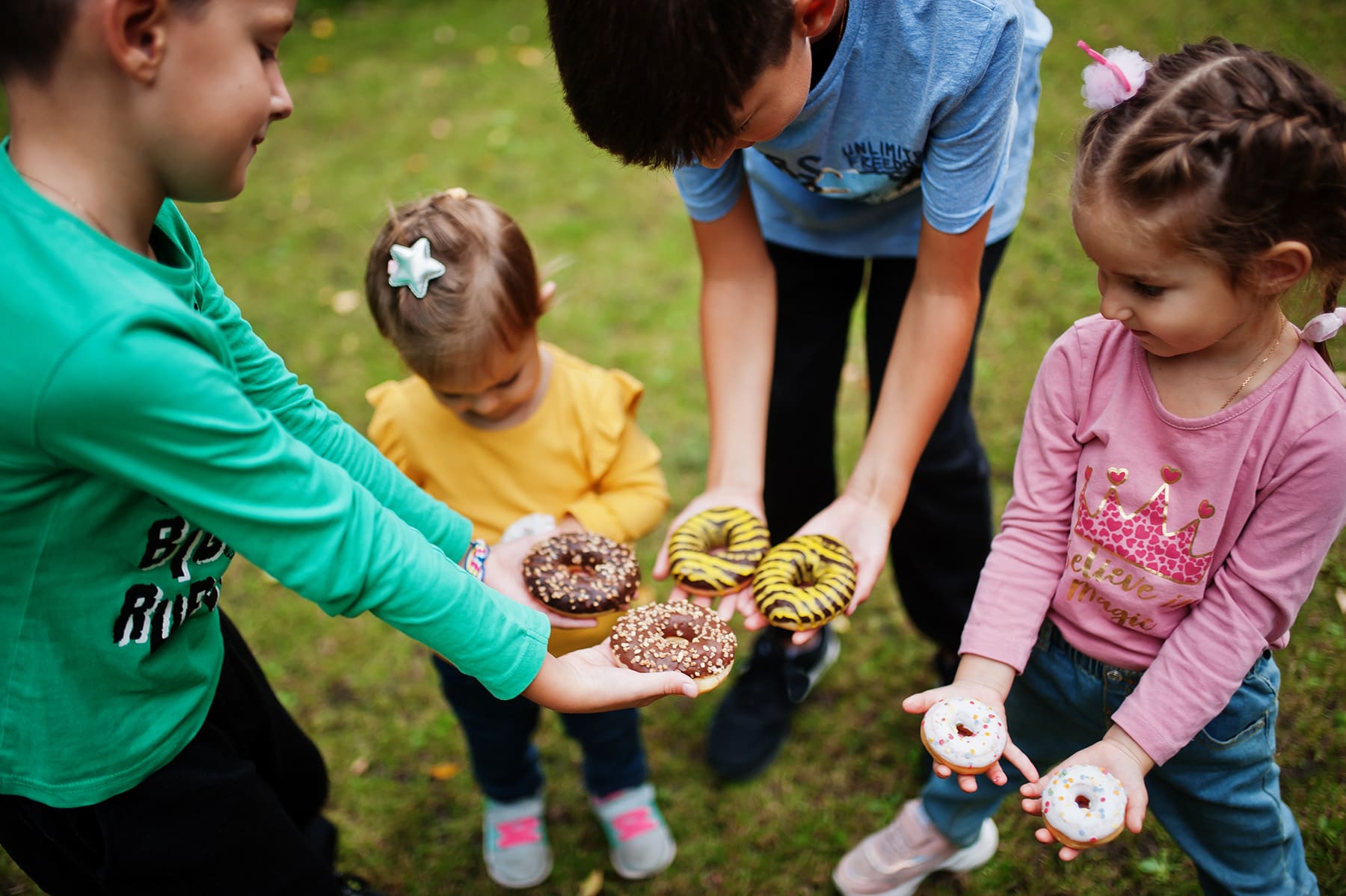Four young children standing outdoors, each holding colorful doughnuts in their hands, sharing treats and enjoying a playful moment in a grassy yard.