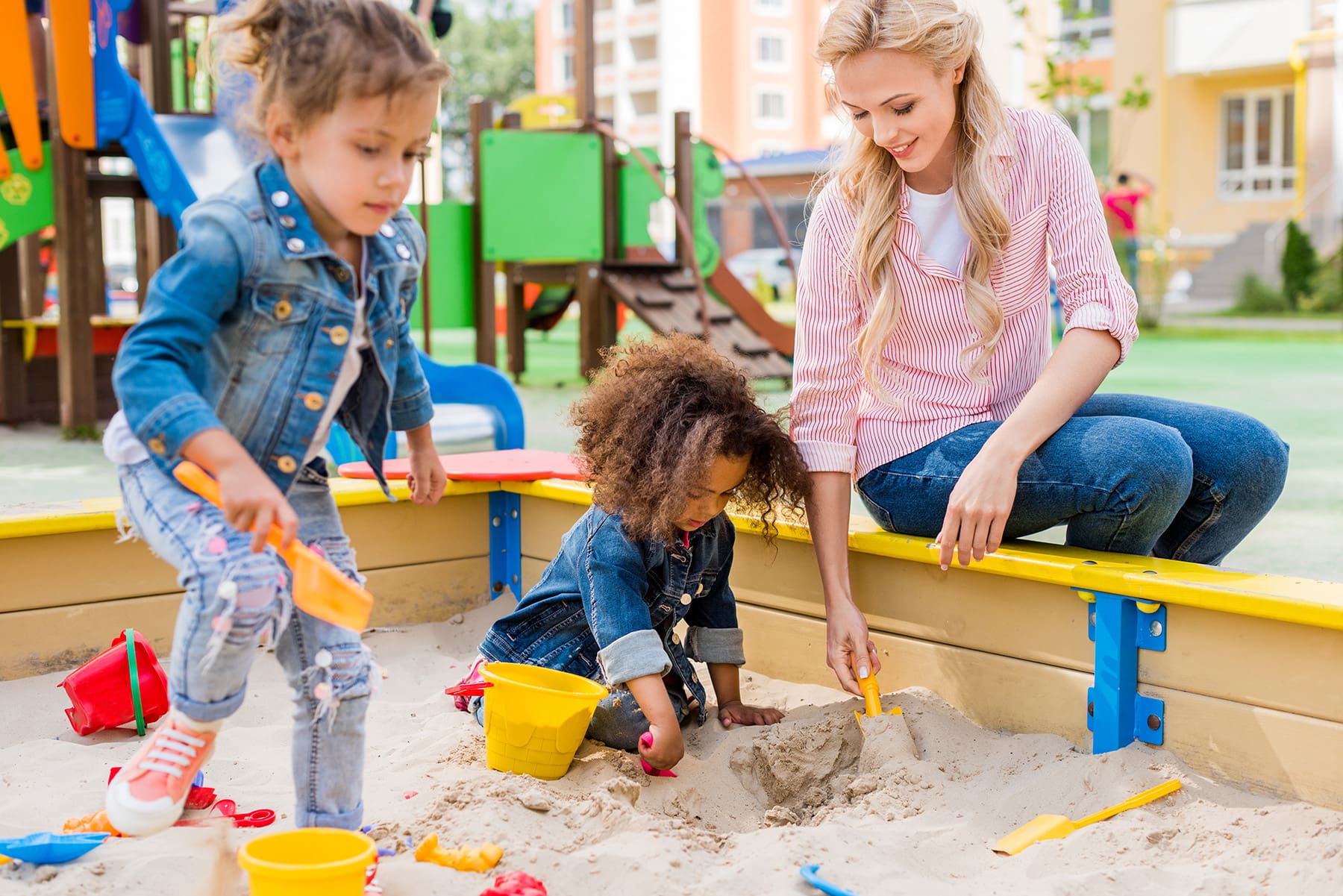 Mother and two young girls playing together in a sandbox at a colorful outdoor playground, using toy shovels and buckets to dig in the sand.