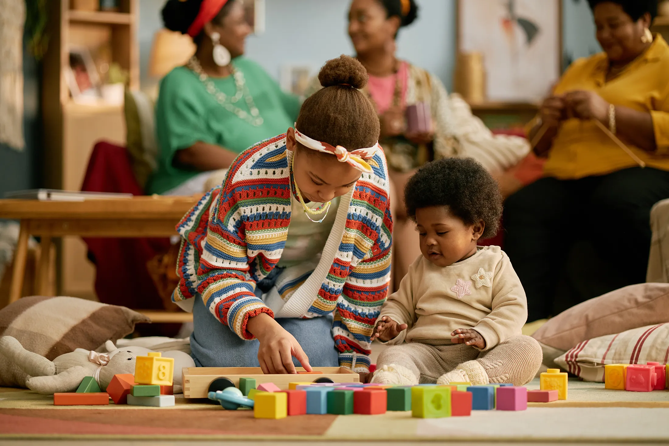 Older sister helping baby sibling play with colorful blocks on the floor while family members watch in a cozy, homey setting.