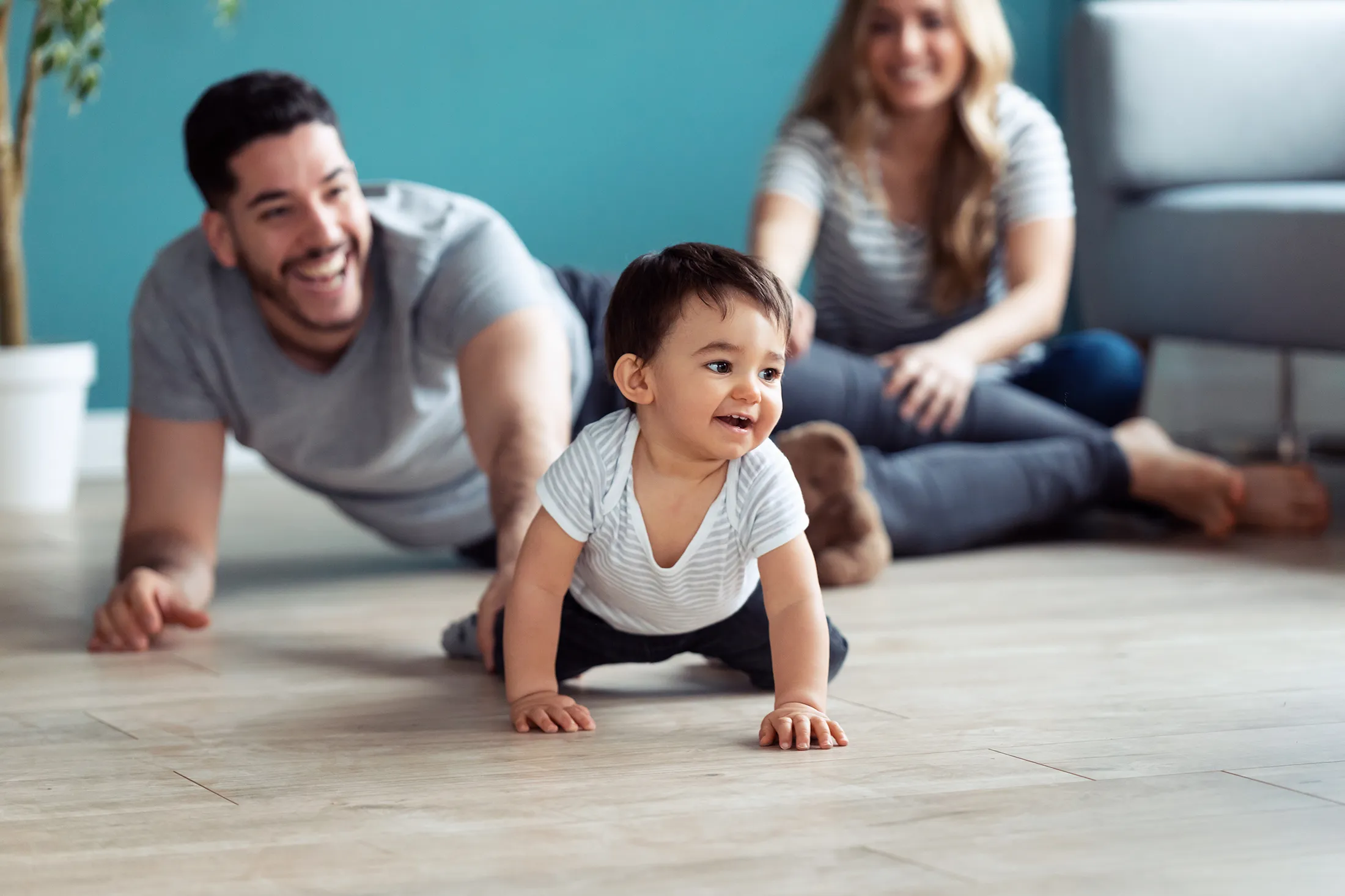 Smiling baby crawling on the floor while parents encourage and play in the background.