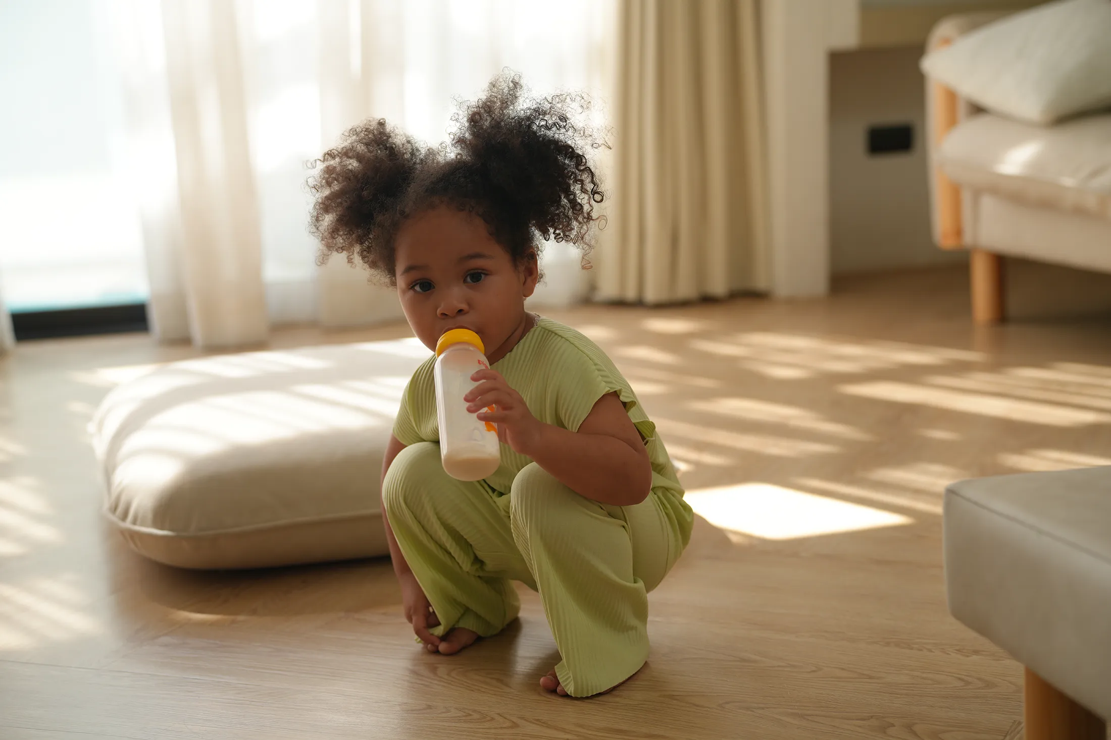 Toddler in green outfit drinking milk from a bottle while sitting on the floor at home.